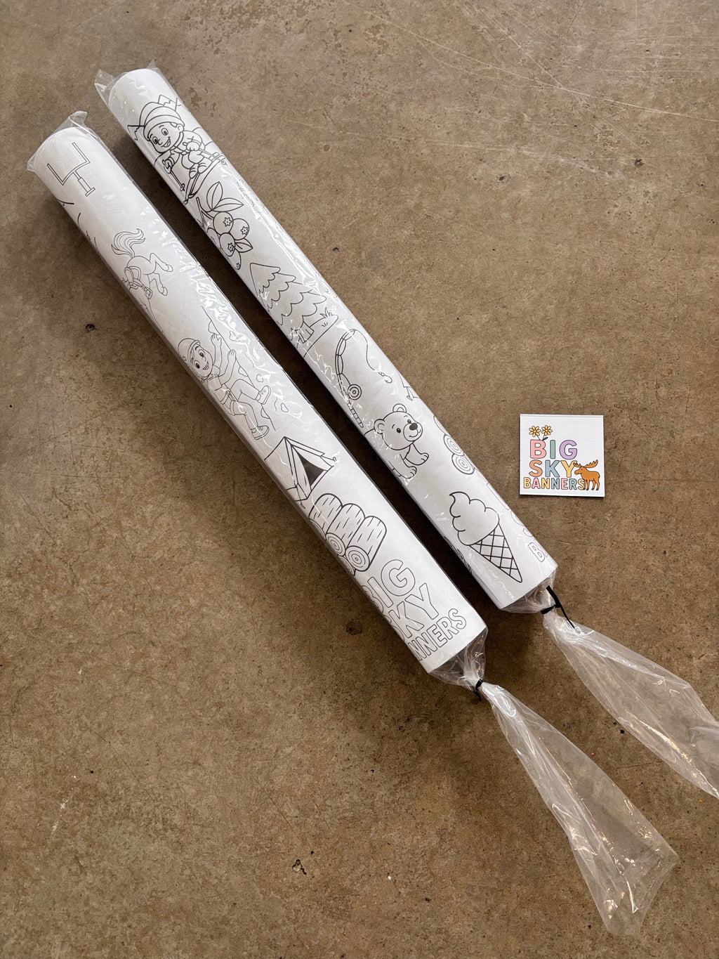 Two white candles with black designs on a brown surface, featuring a small square with text and graphics in the corner.