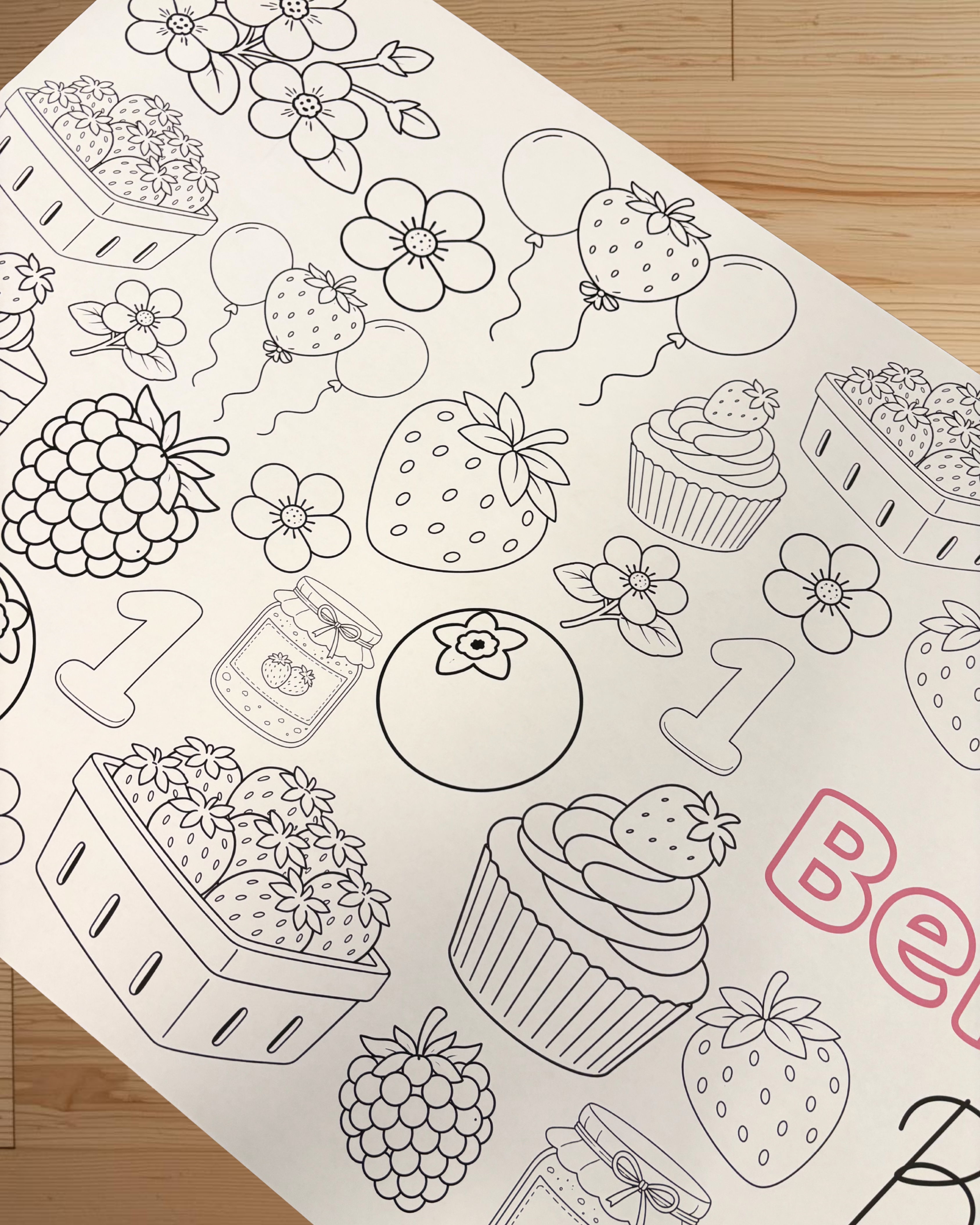 Coloring sheet with black line art of fruits, berries, and a cupcake on a wooden surface.